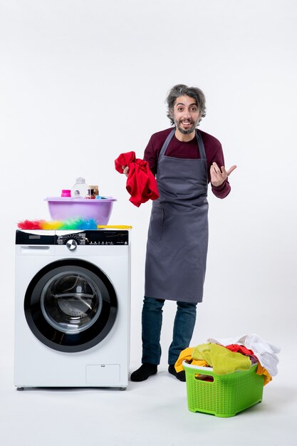 Front view male housekeeper holding red towel standing near washing machine on white wall