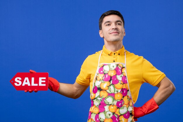 Front view male housekeeper in yellow t-shirt putting hand on his waist holding sale sign on blue space