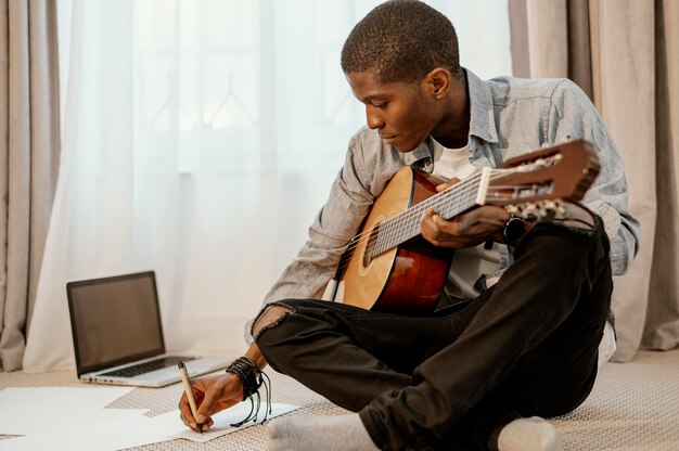 침대와 노트북에 기타와 함께 음악을 쓰는 남성 음악가의 전면보기