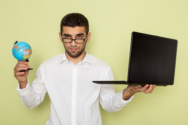 Foto gratuita vista frontale del lavoratore di ufficio maschio in camicia bianca che tiene il suo laptop e piccolo globo sulla parete verde