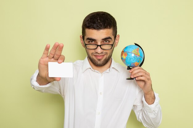 Вид спереди мужского офисного работника в белой рубашке, держащего маленький глобус и карточку на светло-зеленой стене
