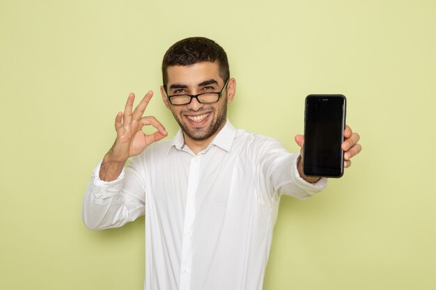 Front view of male office worker in white shirt holding smartphone on the light-green wall