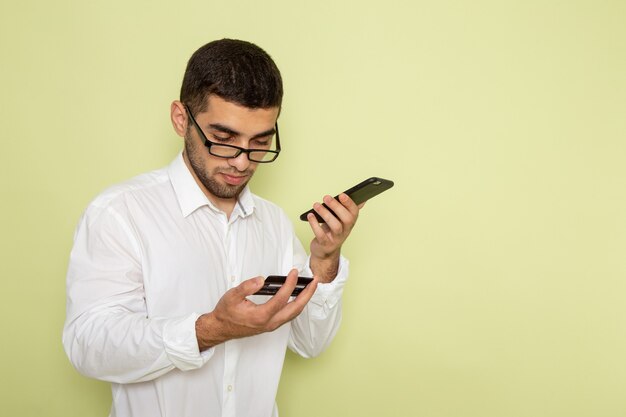Front view of male office worker in white shirt holding and using his phone on the light-green wall