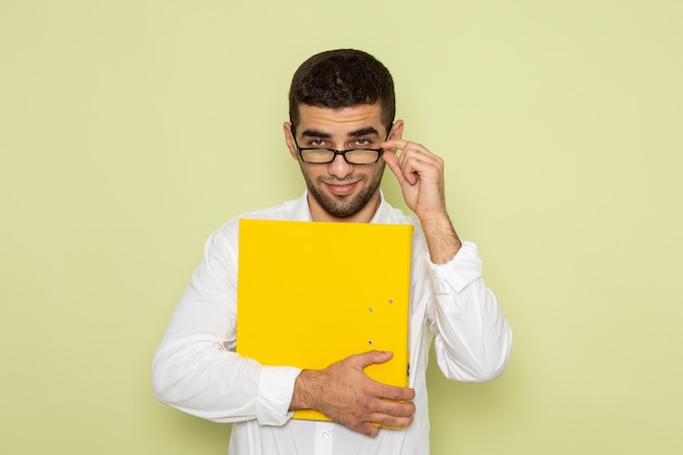 Вид спереди мужского офисного работника в белой рубашке, держащего желтый файл на светло-зеленой стене