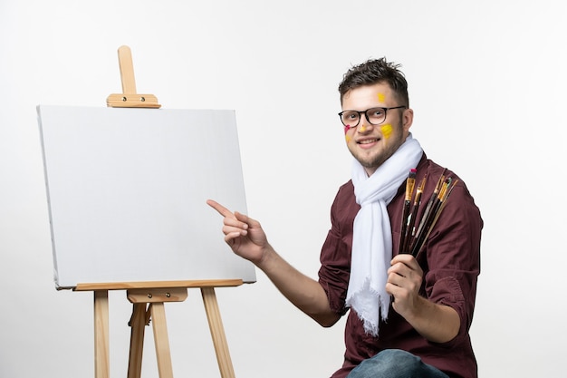 Front view male painter trying to draw on easel holding paint brushes on white wall