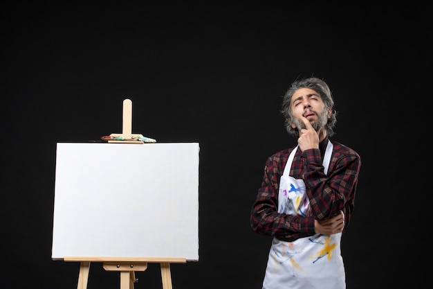 Front view of male painter with easel for drawing standing on black wall