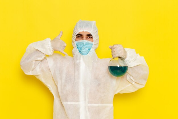 Front view male scientific worker in special protective suit holding flask with blue solution on light-yellow surface