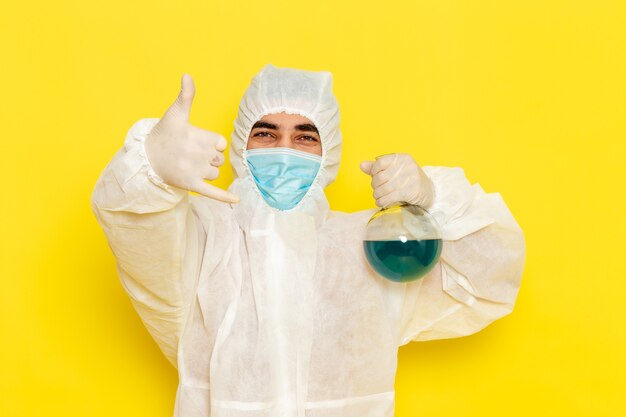 Front view male scientific worker in special protective suit holding flask with blue solution posing on yellow desk