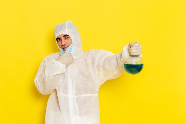 Front view male scientific worker in special protective suit holding flask with blue solution on yellow surface