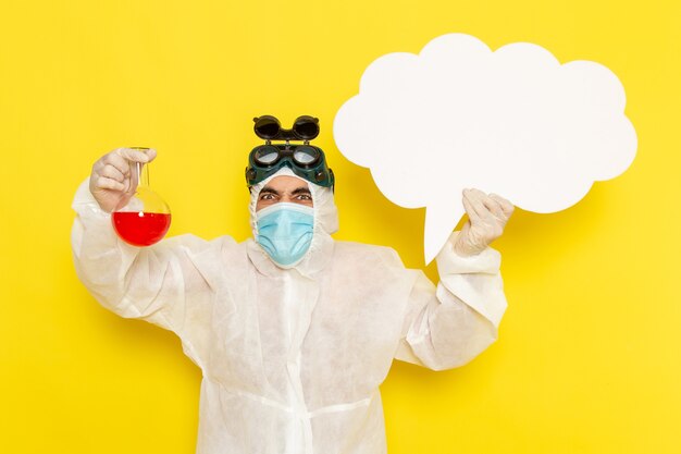 Front view male scientific worker in special protective suit holding flask with red solution and big white sign on the yellow desk