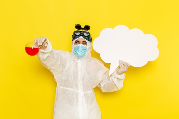 Front view male scientific worker in special protective suit holding flask with red solution and big white sign on yellow desk