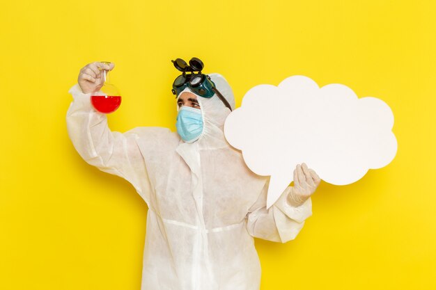 Front view male scientific worker in special protective suit holding flask with red solution and big white sign on the yellow surface