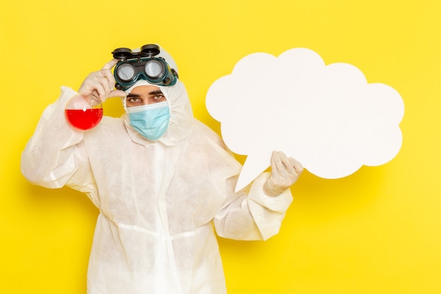 Front view male scientific worker in special protective suit holding flask with red solution and big white sign on yellow surface