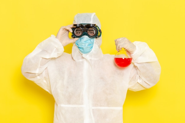 Front view male scientific worker in special protective suit holding flask with red solution on light yellow surface