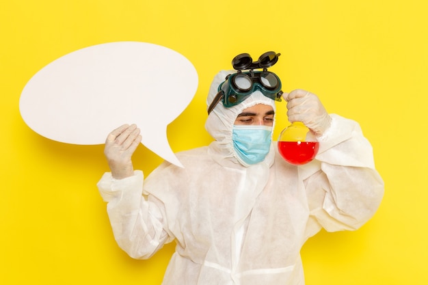 Front view male scientific worker in special protective suit holding flask with red solution and white sign on light-yellow desk