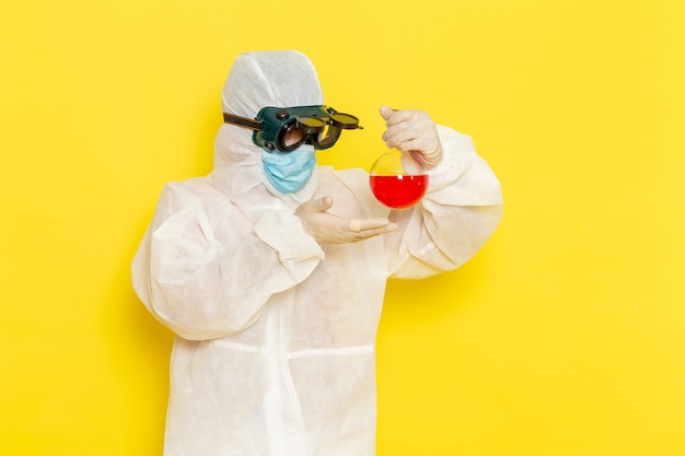 Вид спереди научный сотрудник-мужчина в специальном защитном костюме, держащий фляжку с красным раствором на желтом столе