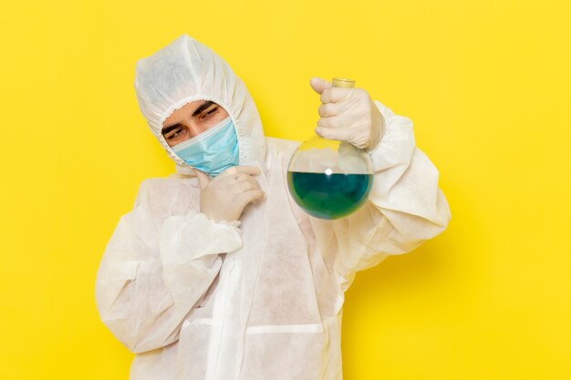 Front view of male scientific worker in special protective suit with mask holding flask and thinking on yellow wall