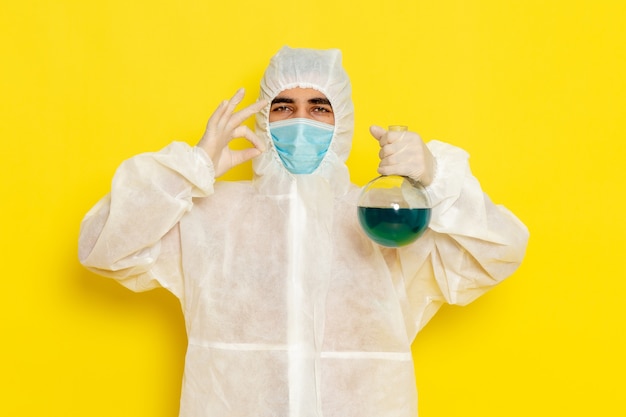 Вид спереди научного работника-мужчины в специальном защитном костюме с маской, держащей фляжку на желтой стене