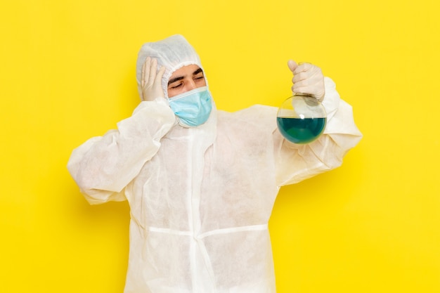 Front view of male scientific worker in special protective suit with mask holding flask on yellow wall