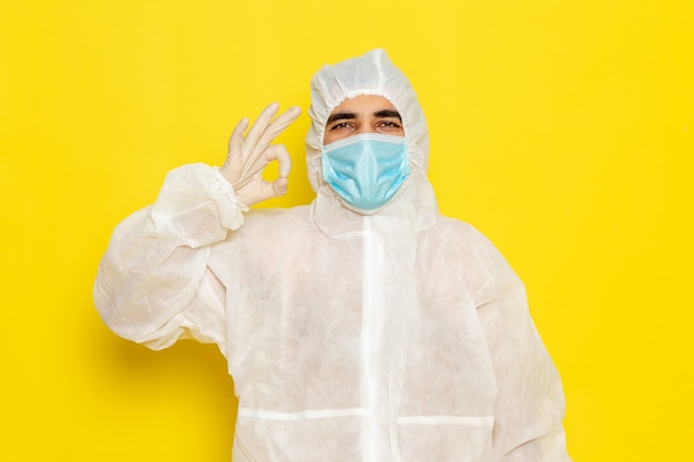 Foto gratuita vista frontale del lavoratore scientifico maschio in vestito bianco protettivo speciale con la maschera che mostra il segno giusto sulla parete gialla