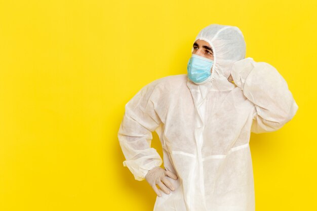 Front view of male scientific worker in special protective white suit and with mask thinking on yellow wall