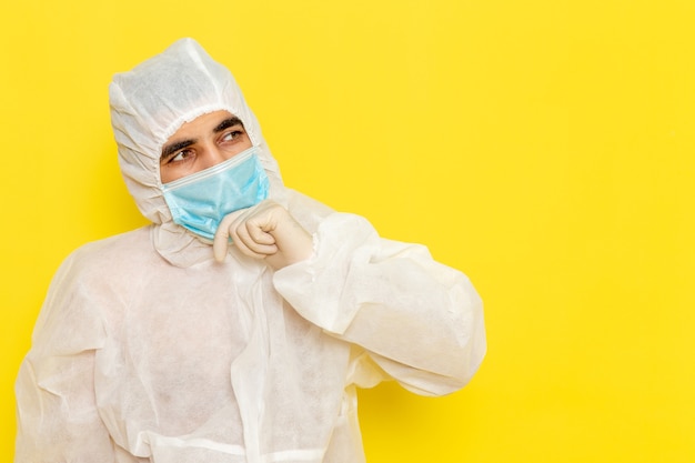 Front view of male scientific worker in special protective white suit with sterile mask just thinking on yellow wall