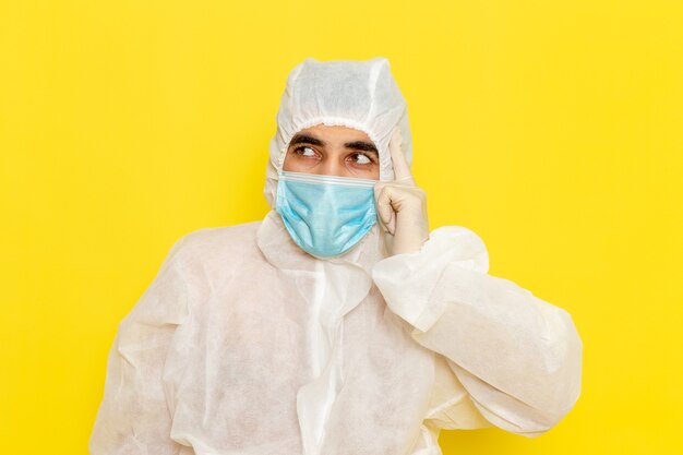 Front view of male scientific worker in special protective white suit with sterile mask thinking on yellow wall