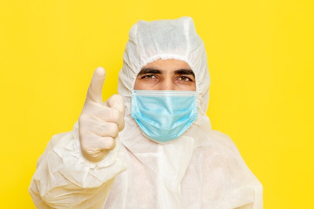 Front view of male scientific worker in special protective white suit with sterile mask threatens on yellow wall