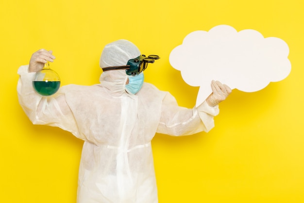 Front view male scientific worker in special suit holding flask with green solution and big white sign on yellow desk