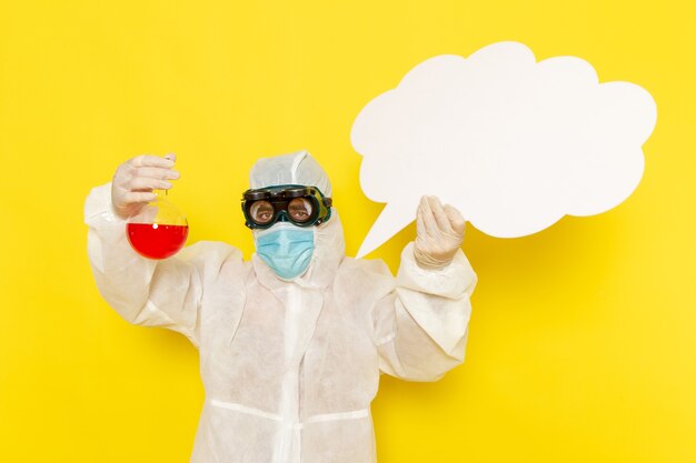 Front view male scientific worker in special suit holding flask with red solution and big white sign on yellow desk