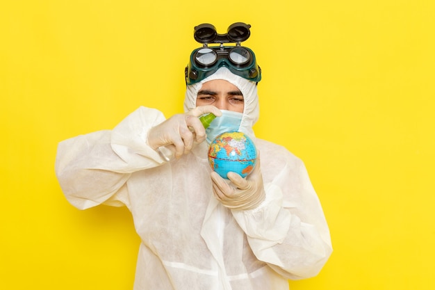 Front view male scientific worker in special suit holding little round globe spraying it on yellow surface
