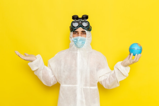 Front view male scientific worker in special suit holding little round globe on the yellow desk