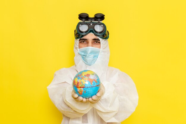 Front view male scientific worker in special suit holding little round globe on yellow floor