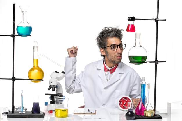 Вид спереди ученый-мужчина в медицинском костюме, сидящий перед столом с растворами на светлом белом фоне, вирусная химия здоровья covid- lab