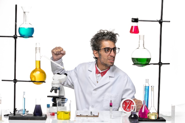 Front view male scientist in medical suit sitting in front of table with solutions on a white background covid lab virus health chemistry