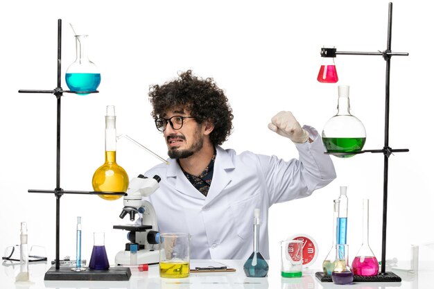 Front view male scientist in medical suit sitting with solutions and microscope on light white space