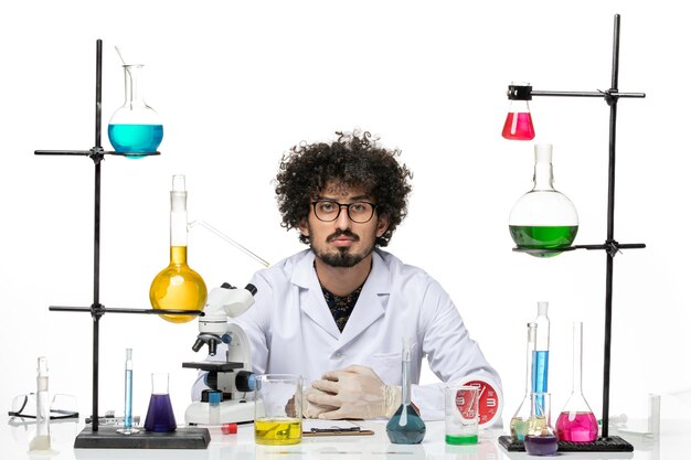 Front view male scientist in medical suit sitting with solutions and microscope on a white space