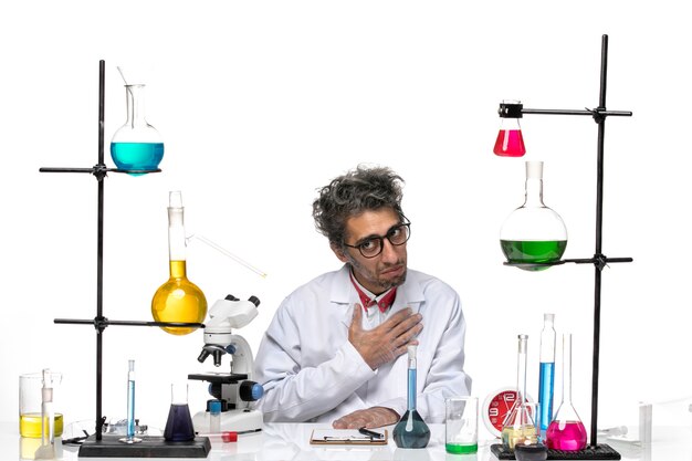 Вид спереди ученый-мужчина в белом медицинском костюме, сидящий перед столом с растворами