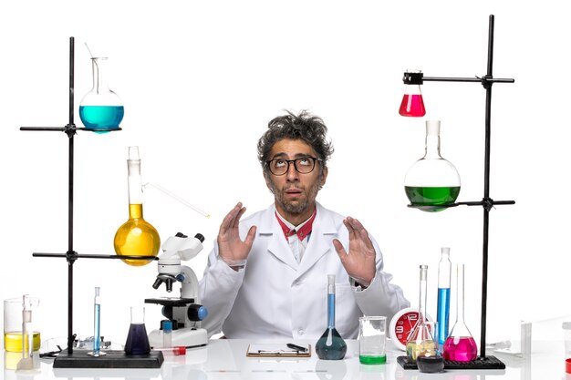 Front view male scientist in white medical suit sitting in front of table with solutions