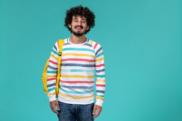 Front view of male student in colored striped shirt wearing yellow backpack on blue wall
