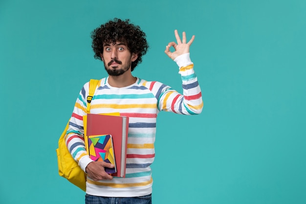 Front view of male student in colored striped shirt wearing yellow backpack holding files and copybooks on blue wall