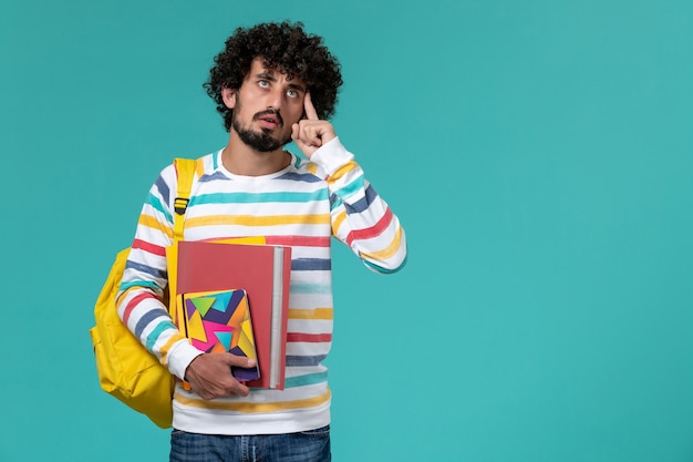 Front view of male student in colored striped shirt wearing yellow backpack holding files and copybooks thinking on blue wall