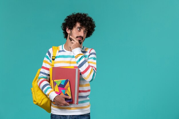 Front view of male student in colored striped shirt wearing yellow backpack holding files and copybooks thinking on blue wall