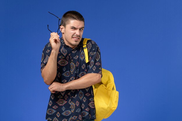 Front view of male student in dark cotton shirt wearing yellow backpack posing on light blue wall