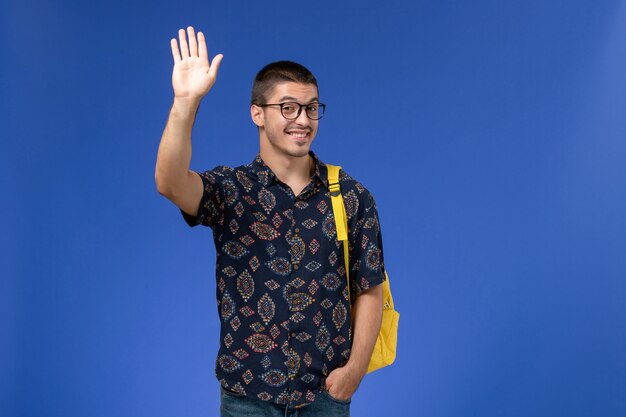 Front view of male student in dark cotton shirt wearing yellow backpack waving his hand on blue wall