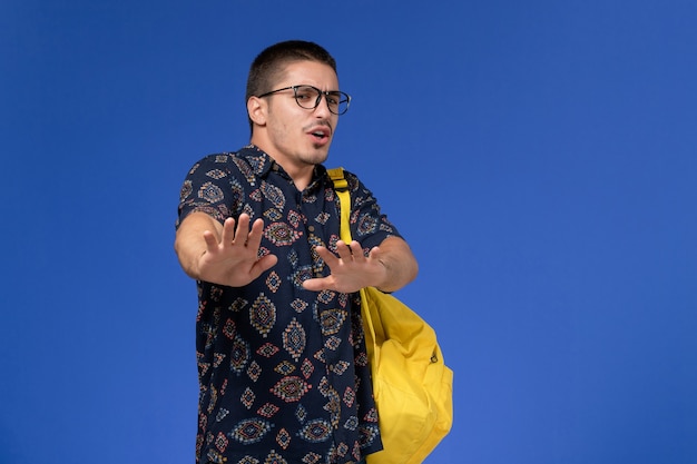 Free photo front view of male student in dark cotton shirt wearing yellow backpack wearing optical sunglasses posing on the blue wall