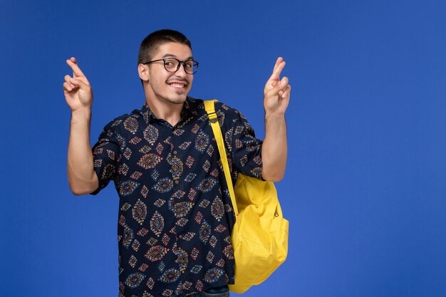 Front view of male student in dark shirt wearing yellow backpac crossing his fingersk on the blue wall