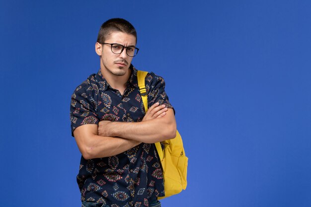 Front view of male student in dark shirt wearing yellow backpack posing on blue wall