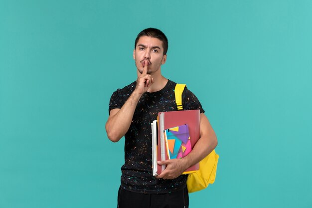 Front view of male student in dark t-shirt wearing yellow backpack holding copybook and files on blue wall