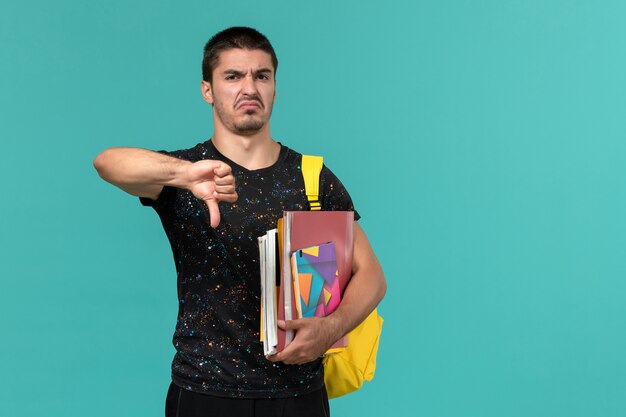 Front view of male student in dark t-shirt wearing yellow backpack holding copybook and files on blue wall